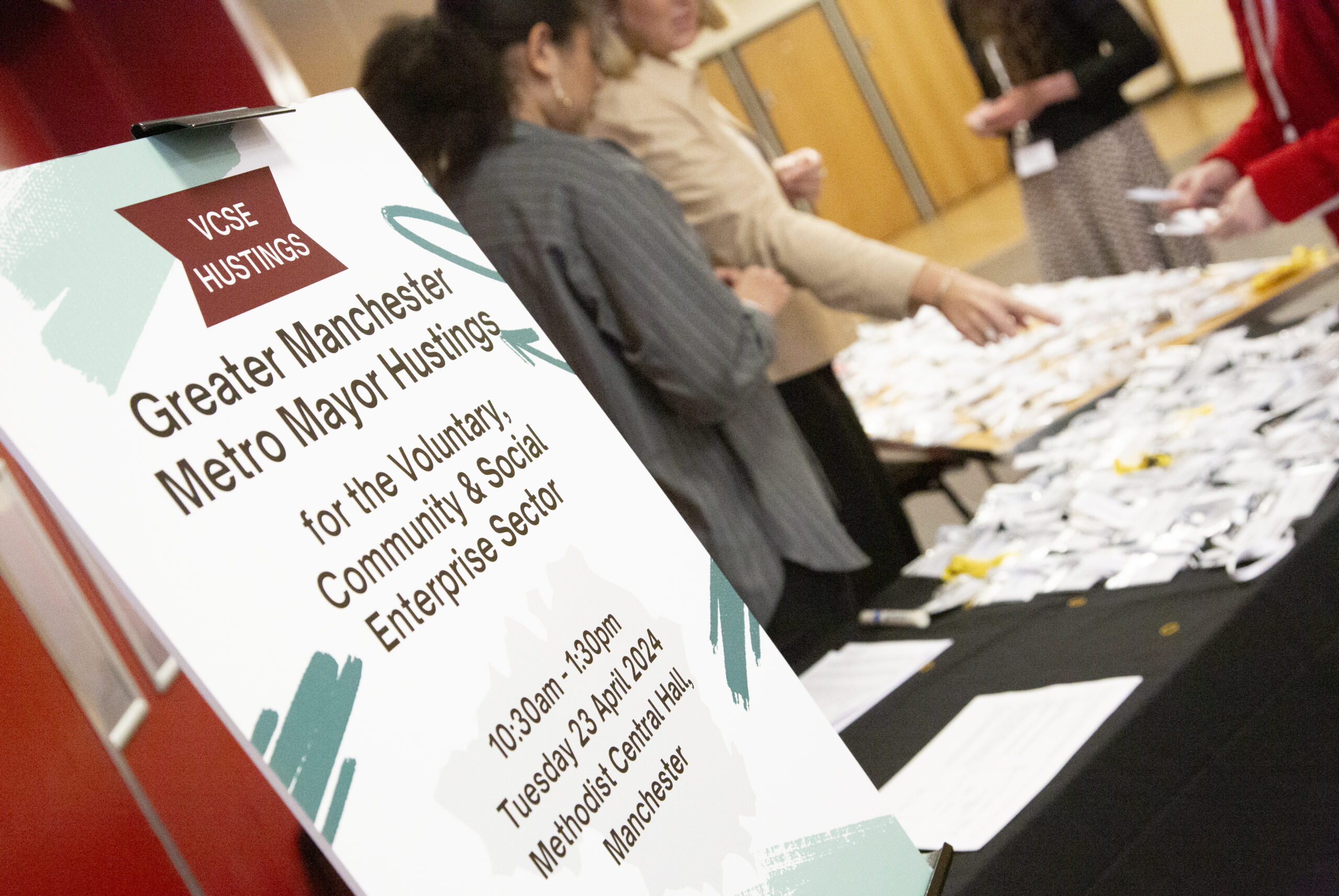 Photograph of the arrival desk for the GM VCSE hustings event that took place last week. There is a easel with the event poster on it, next to two staff members laying out name badges for attendees.