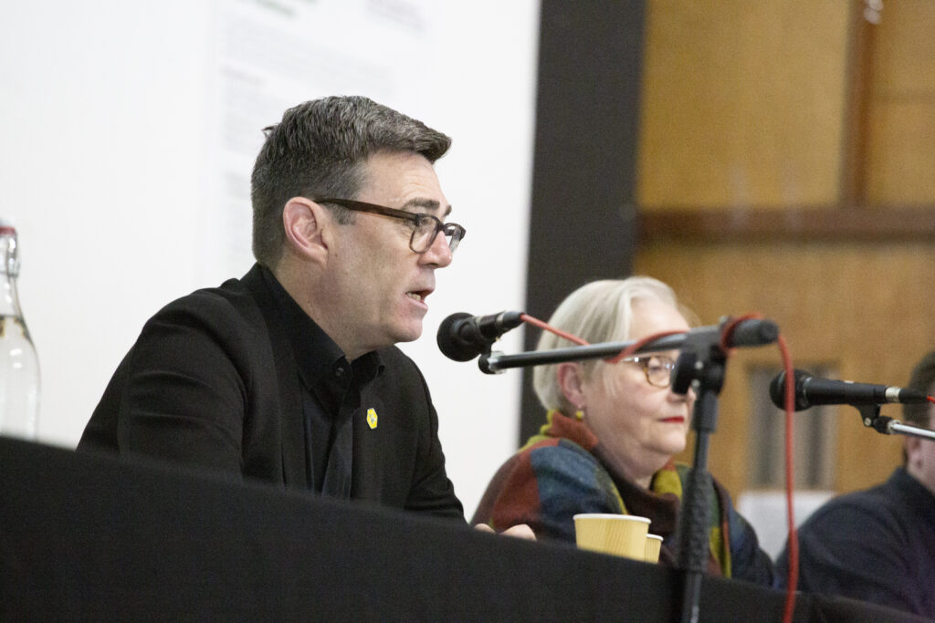 Photo of Andy Burnham sitting and talking a microphone on stage behind a table with a black tablecloth. Astrid Johnson, representing Green is in the background, out of focus.